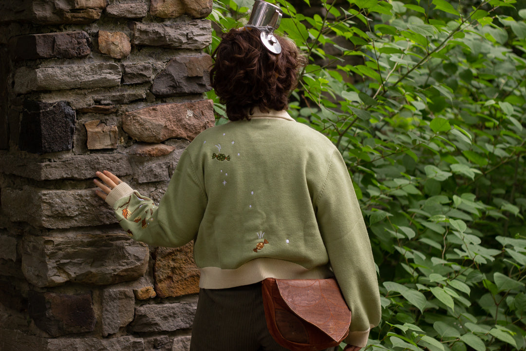 A soft, button-up, cropped, knit cardigan, in shades of soft sage and warm cream. The cardigan features whimsical embroidered details of autumn leaves, candy, snow covered branches, teapot, and froggy friend.