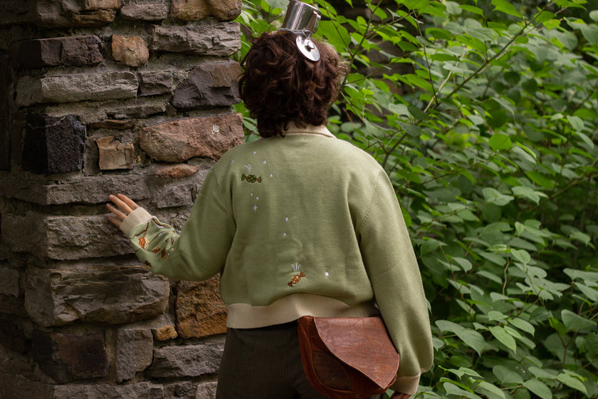 A soft, button-up, cropped, knit cardigan, in shades of soft sage and warm cream. The cardigan features whimsical embroidered details of autumn leaves, candy, snow covered branches, teapot, and froggy friend.