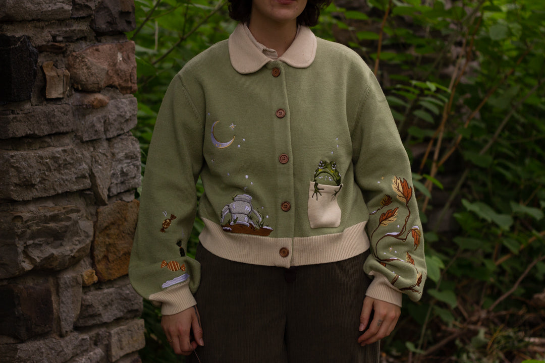 A soft, button-up, cropped, knit cardigan, in shades of soft sage and warm cream. The cardigan features whimsical embroidered details of autumn leaves, candy, snow covered branches, teapot, and froggy friend.