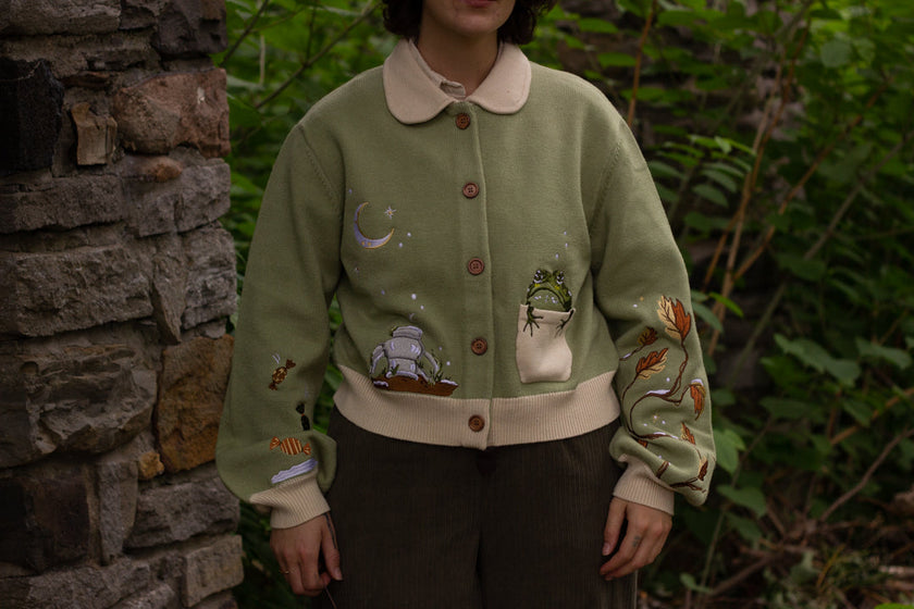 A soft, button-up, cropped, knit cardigan, in shades of soft sage and warm cream. The cardigan features whimsical embroidered details of autumn leaves, candy, snow covered branches, teapot, and froggy friend.