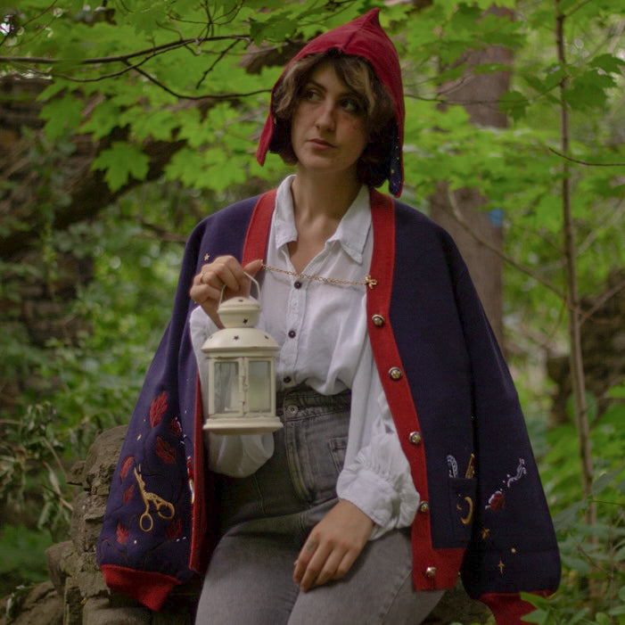 A soft, button-up, oversized, knit cardigan, in shades of navy blue, crimson red, and vintage gold. The cardigan features whimsical embroidered details of autumn leaves, moon and stars, a snowy lantern, and a bluebirds feather.