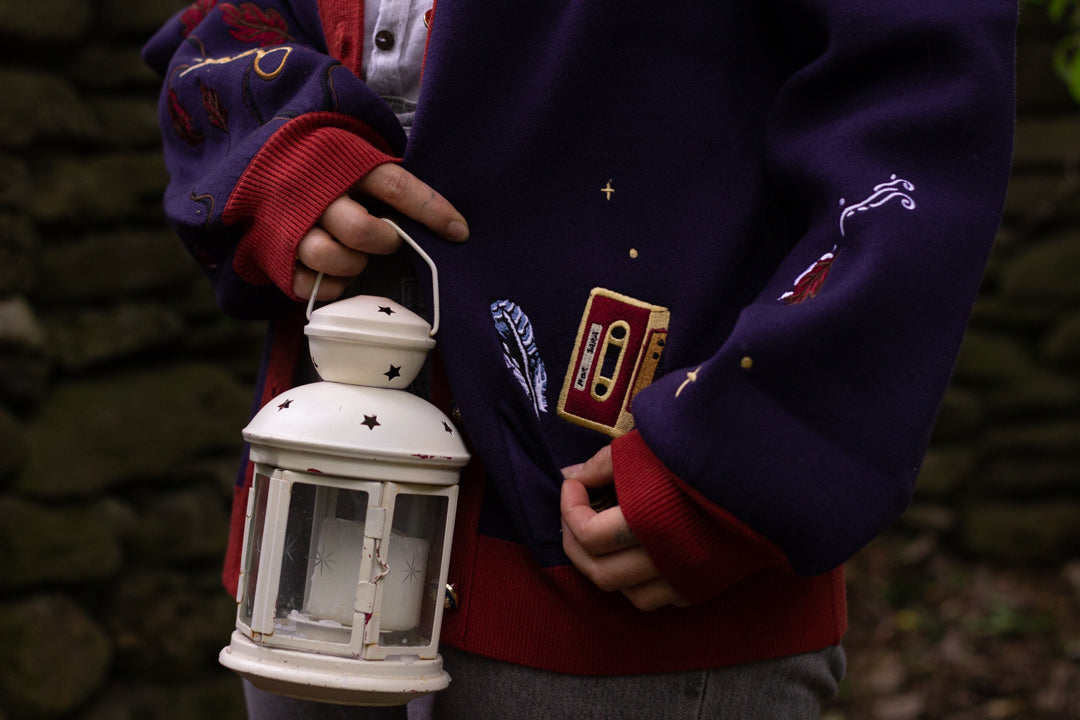 A soft, button-up, oversized, knit cardigan, in shades of navy blue, crimson red, and vintage gold. The cardigan features whimsical embroidered details of autumn leaves, moon and stars, a snowy lantern, and a bluebirds feather.