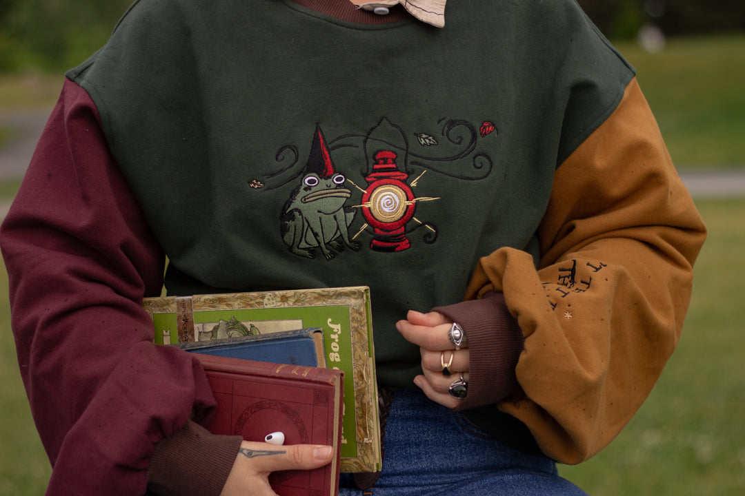 Cozy, oversized, puff-sleeved, color-blocked, cotton crewneck sweater in forest green, with burgundy, and mustard coloured sleeves. The sweater features an intricately embroidered design of a green frog with a red wizard hat and a red lantern on the chest.