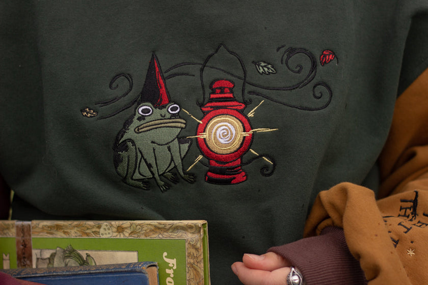Cozy, oversized, puff-sleeved, color-blocked, cotton crewneck sweater in forest green, with burgundy, and mustard coloured sleeves. The sweater features an intricately embroidered design of a green frog with a red wizard hat and a red lantern on the chest.
