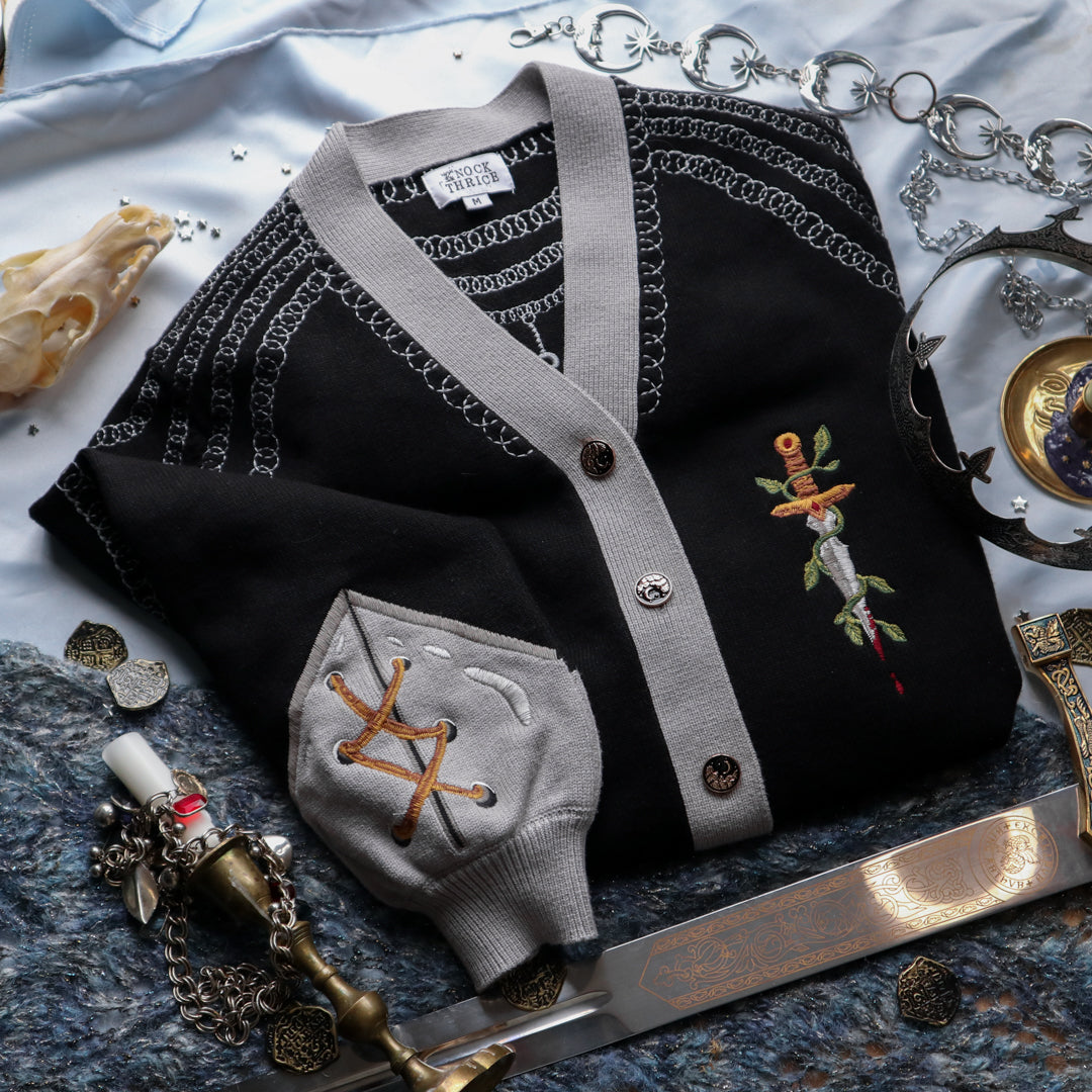 A soft button-up black and silver knit cardigan. With black and gold moon buttons, the design features intricate embroidery of a vine wrapped sword, chainmail, and armour. Evoking a whimsical and fantasy-inspired look.