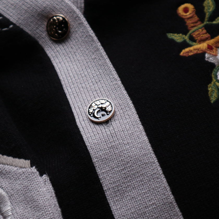 A soft button-up black and silver knit cardigan. With black and gold moon buttons, the design features intricate embroidery of a vine wrapped sword, chainmail, and armour. Evoking a whimsical and fantasy-inspired look.