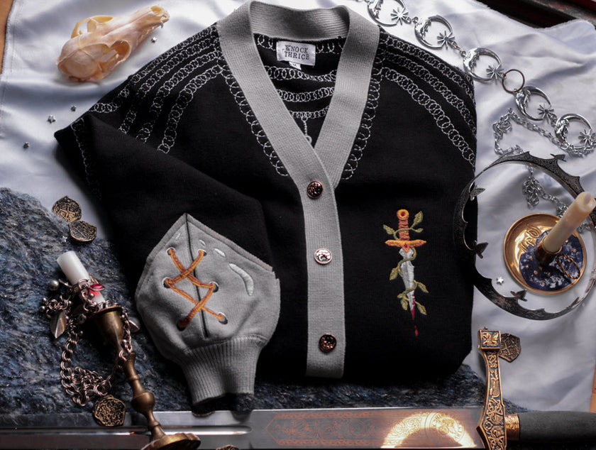 A soft button-up black and silver knit cardigan. With black and gold moon buttons, the design features intricate embroidery of a vine wrapped sword, chainmail, and armour. Evoking a whimsical and fantasy-inspired look.