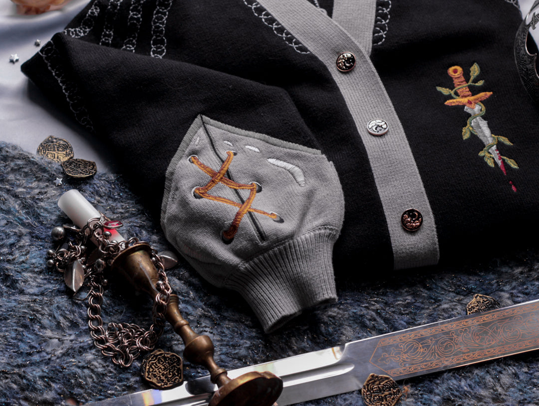 A soft button-up black and silver knit cardigan. With black and gold moon buttons, the design features intricate embroidery of a vine wrapped sword, chainmail, and armour. Evoking a whimsical and fantasy-inspired look.