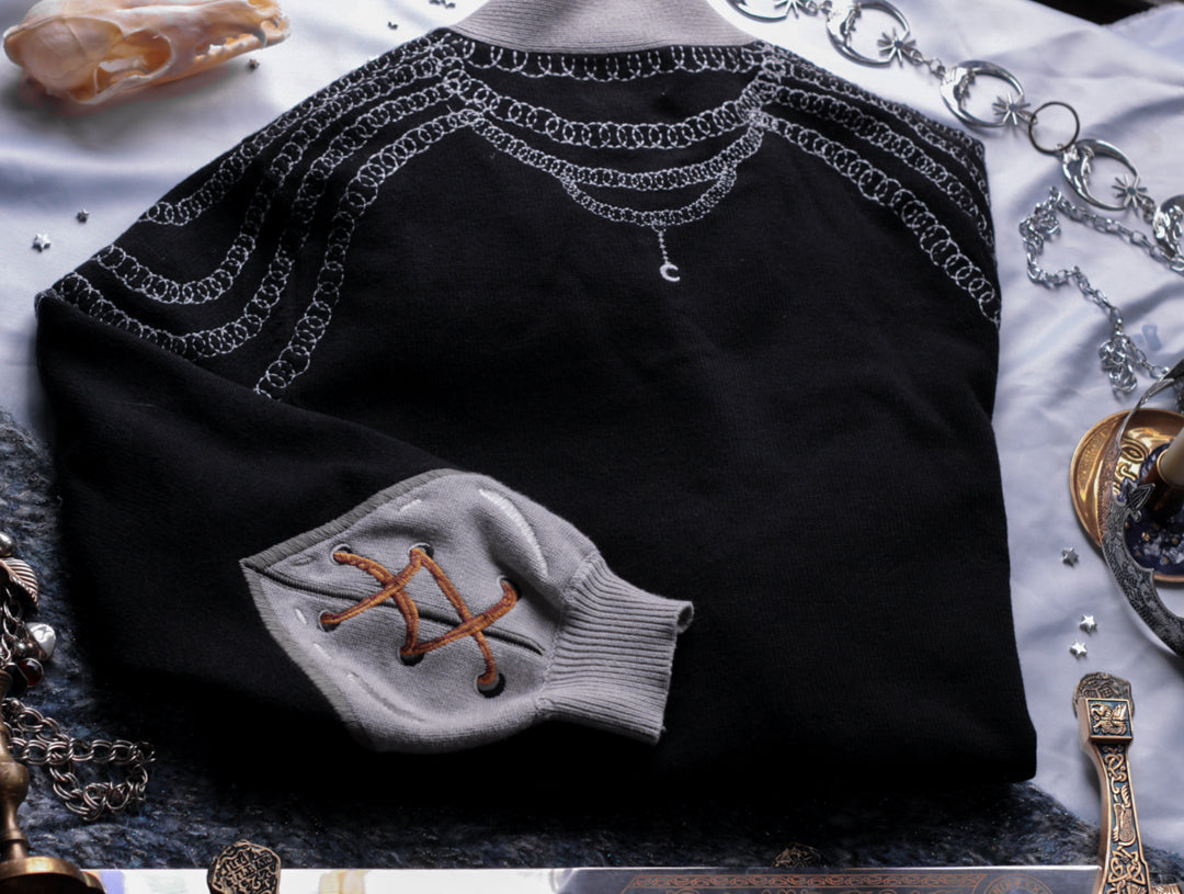 A soft button-up black and silver knit cardigan. With black and gold moon buttons, the design features intricate embroidery of a vine wrapped sword, chainmail, and armour. Evoking a whimsical and fantasy-inspired look.