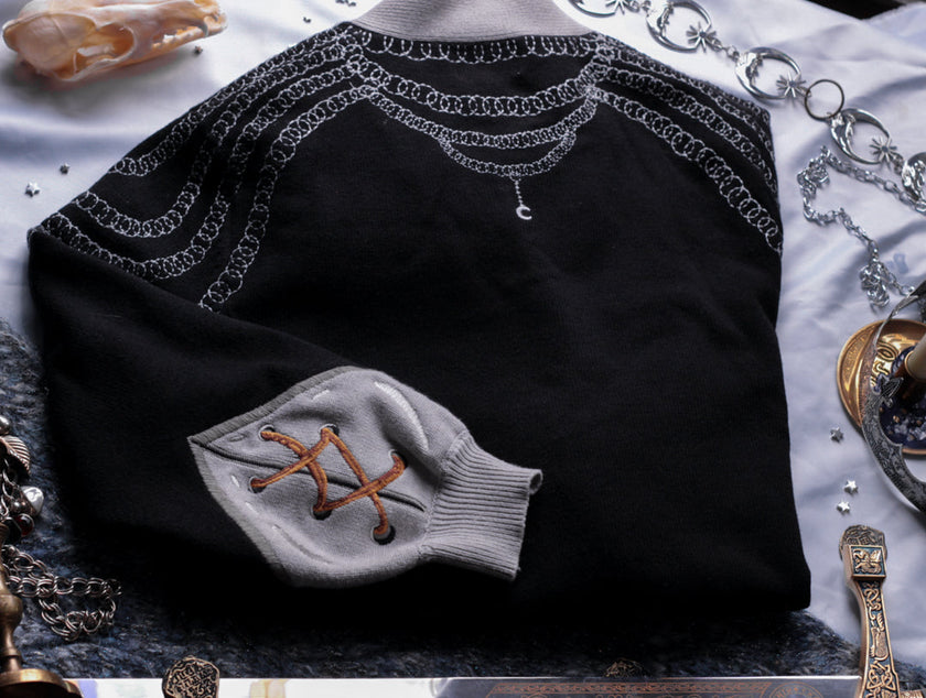 A soft button-up black and silver knit cardigan. With black and gold moon buttons, the design features intricate embroidery of a vine wrapped sword, chainmail, and armour. Evoking a whimsical and fantasy-inspired look.