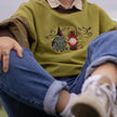 Cozy, oversized, puff-sleeved, color-blocked, cotton crewneck sweater in swamp-green, and light and dark brown. The sweater features an intricately embroidered design of a green frog with a red wizard hat and a red lantern on the chest.