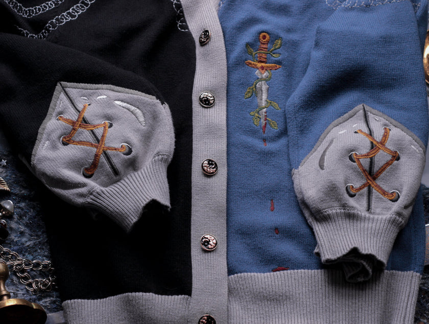 A soft button-up black and silver knit cardigan. With black and gold moon buttons, the design features intricate embroidery of a vine wrapped sword, chainmail, and armour. Evoking a whimsical and fantasy-inspired look.
