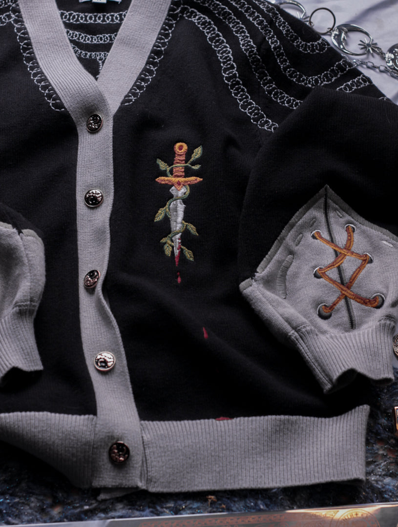A soft button-up black and silver knit cardigan. With black and gold moon buttons, the design features intricate embroidery of a vine wrapped sword, chainmail, and armour. Evoking a whimsical and fantasy-inspired look.