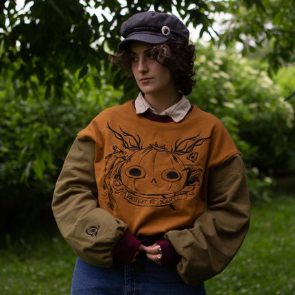 Cozy, oversized, puff-sleeved, color-blocked, cotton crewneck sweater in pumpkin-orange, olive, and burgundy. The sweater features an intricately embroidered design of a pumpkin head, branches, and other autumnal elements.