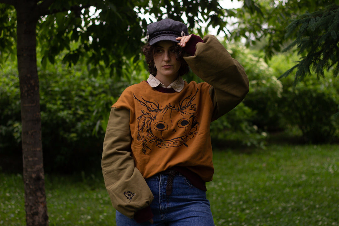 Cozy, oversized, puff-sleeved, color-blocked, cotton crewneck sweater in pumpkin-orange, olive, and burgundy. The sweater features an intricately embroidered design of a pumpkin head, branches, and other autumnal elements.