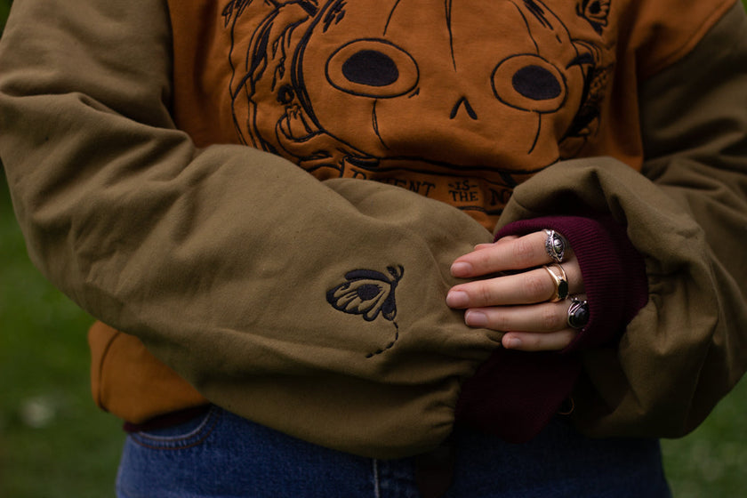 Cozy, oversized, puff-sleeved, color-blocked, cotton crewneck sweater in pumpkin-orange, olive, and burgundy. The sweater features an intricately embroidered design of a pumpkin head, branches, and other autumnal elements.