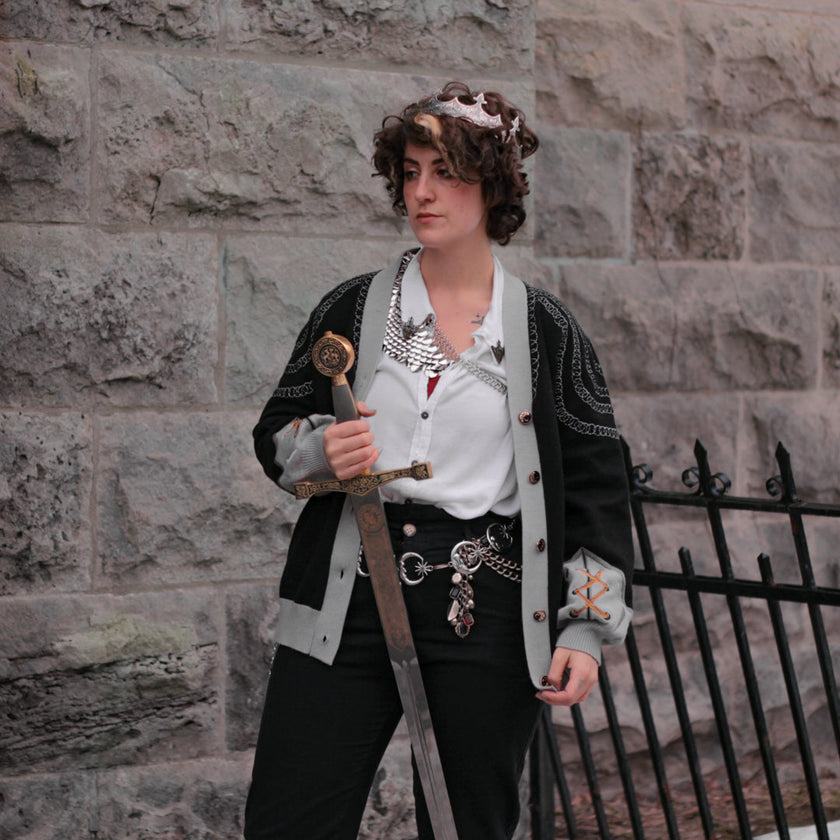 A soft button-up black and silver knit cardigan. With black and gold moon buttons, the design features intricate embroidery of a vine wrapped sword, chainmail, and armour. Evoking a whimsical and fantasy-inspired look.