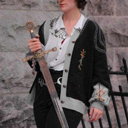 A soft button-up black and silver knit cardigan. With black and gold moon buttons, the design features intricate embroidery of a vine wrapped sword, chainmail, and armour. Evoking a whimsical and fantasy-inspired look.