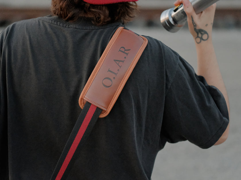Knock Thrice x Rusty Quill canvas satchel with leather straps, plaid side panels, and an embroidered crest design. The bag is lined in plaid and includes a padded laptop pocket, adjustable cross-body strap, and magnetic closure.