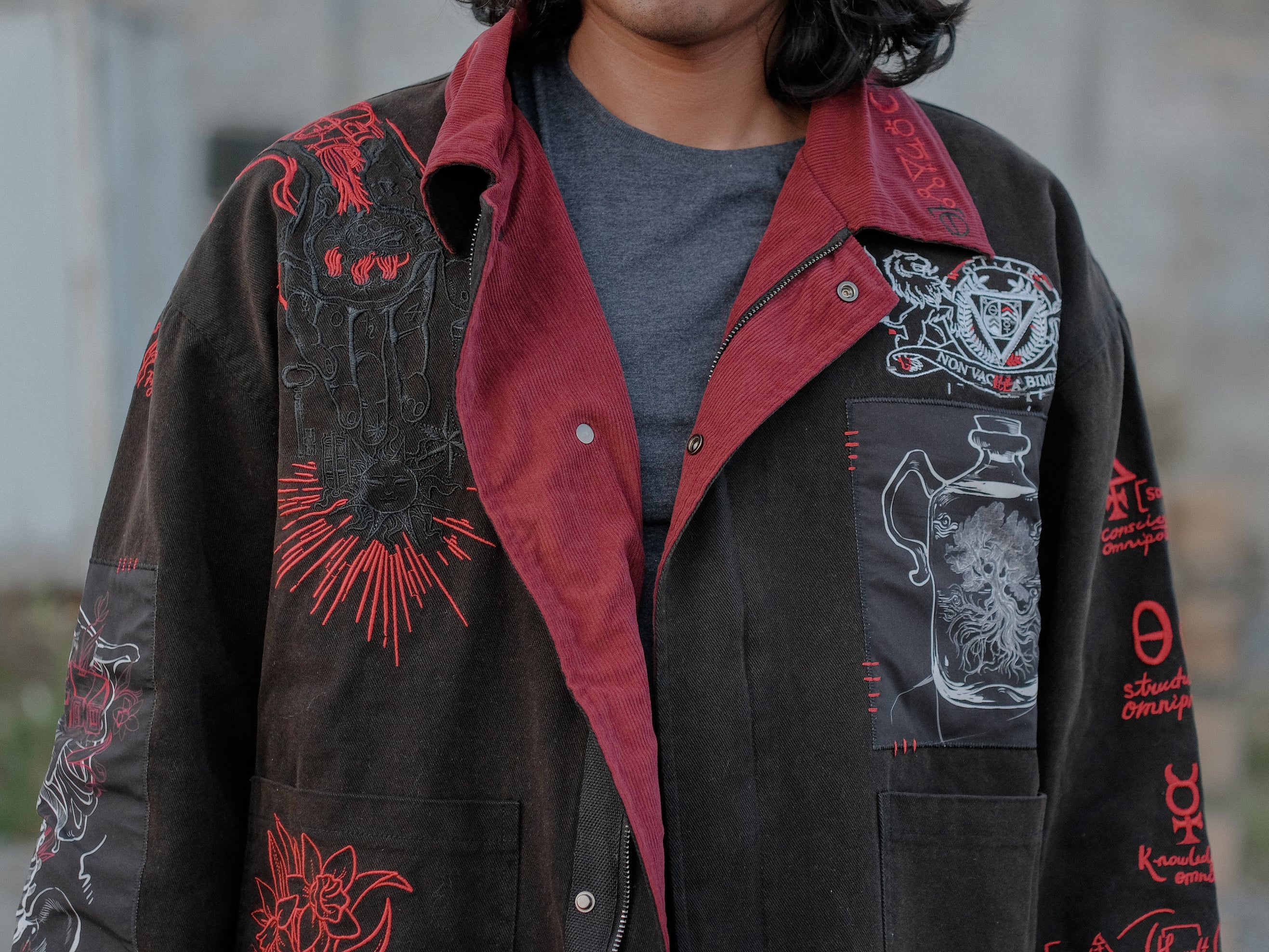 Knock Thrice x Rusty Quill, black fall/spring button-up cotton jacket, with a red corduroy lining. The jacket features both embroidered and patchwork OIAR themed elements, in red, white, and black.