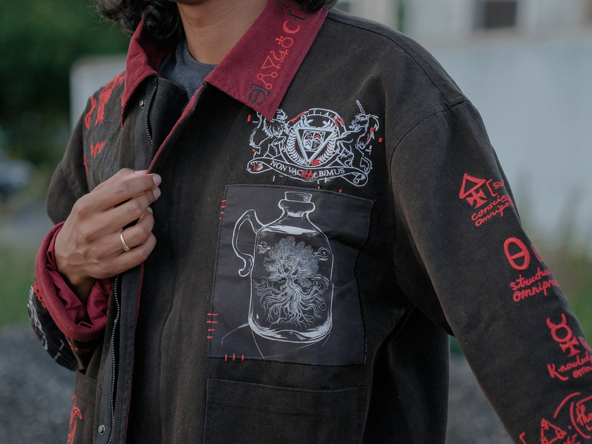 Knock Thrice x Rusty Quill, black fall/spring button-up cotton jacket, with a red corduroy lining. The jacket features both embroidered and patchwork OIAR themed elements, in red, white, and black.