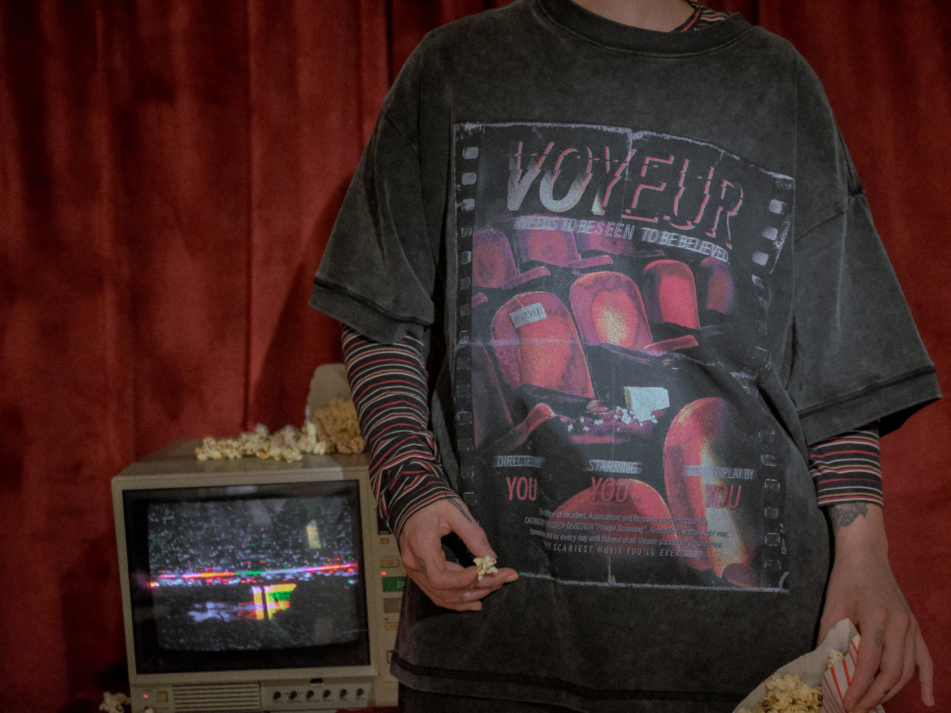 Knock Thrice x Rusty Quill, oversized, stone-washed cotton tee, with a graphic design titled 'Voyeur,' showcasing an eerie movie theatre with red seats, bold text reading 'Needs to be seen to be believed,' and film-inspired details.