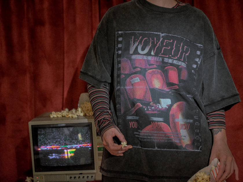 Knock Thrice x Rusty Quill, oversized, stone-washed cotton tee, with a graphic design titled 'Voyeur,' showcasing an eerie movie theatre with red seats, bold text reading 'Needs to be seen to be believed,' and film-inspired details.