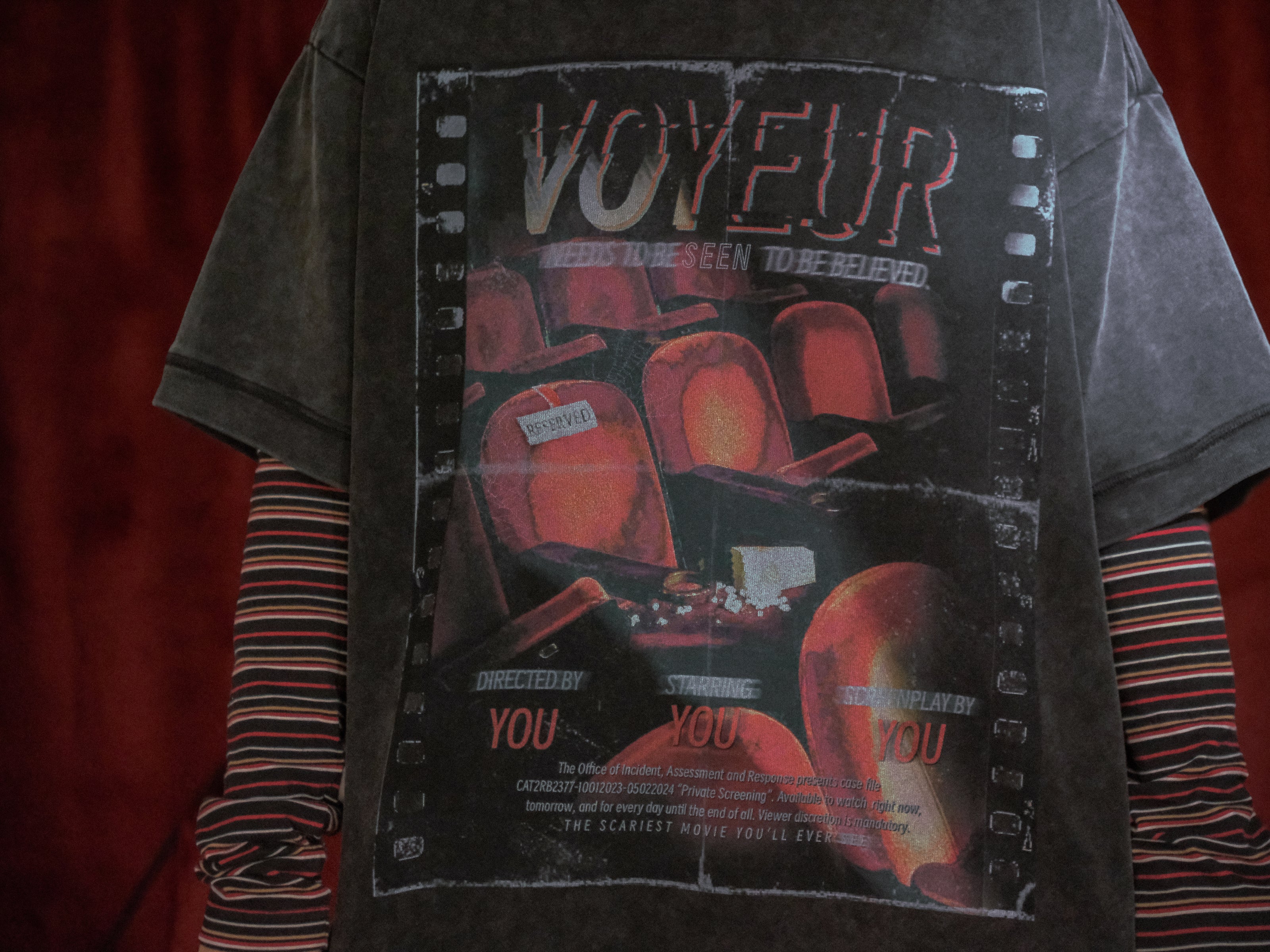 Knock Thrice x Rusty Quill, oversized, stone-washed cotton tee, with a graphic design titled 'Voyeur,' showcasing an eerie movie theatre with red seats, bold text reading 'Needs to be seen to be believed,' and film-inspired details.