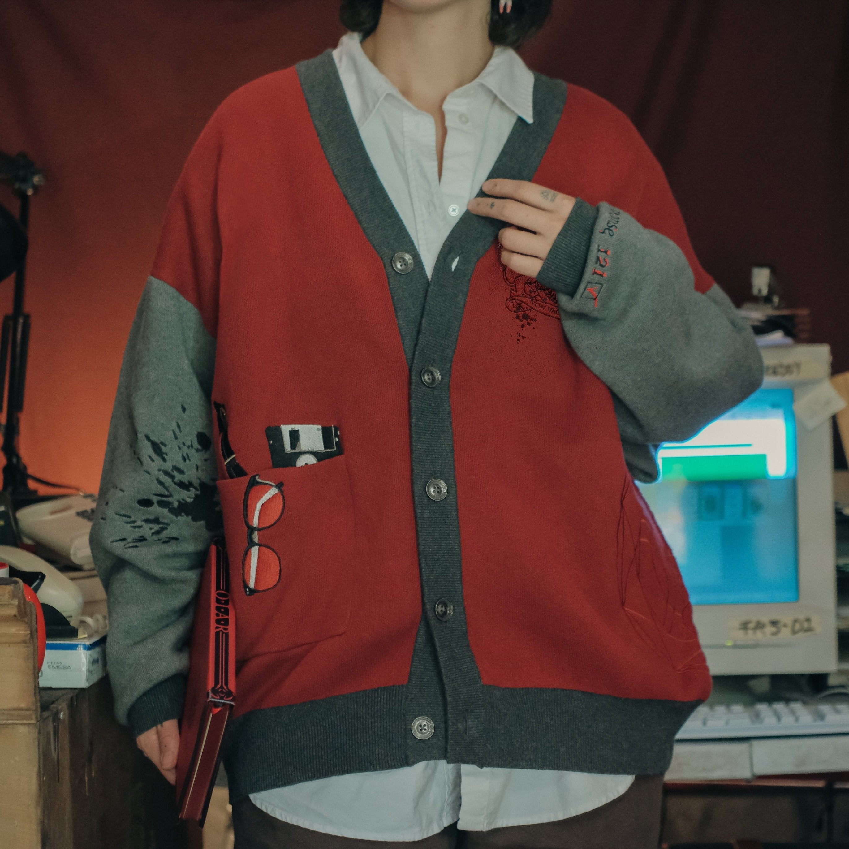 Magnus Protocol, oversized, knit-cardigan, in red, black, and shades of grey. The cardigan features embroidered details of reading glasses, fountain pen, OIAR crest, and reads â€œOffice of Incident Assessment & Responseâ€.