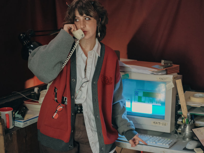 Magnus Protocol, oversized, knit-cardigan, in red, black, and shades of grey. The cardigan features embroidered details of reading glasses, fountain pen, OIAR crest, and reads â€œOffice of Incident Assessment & Responseâ€.