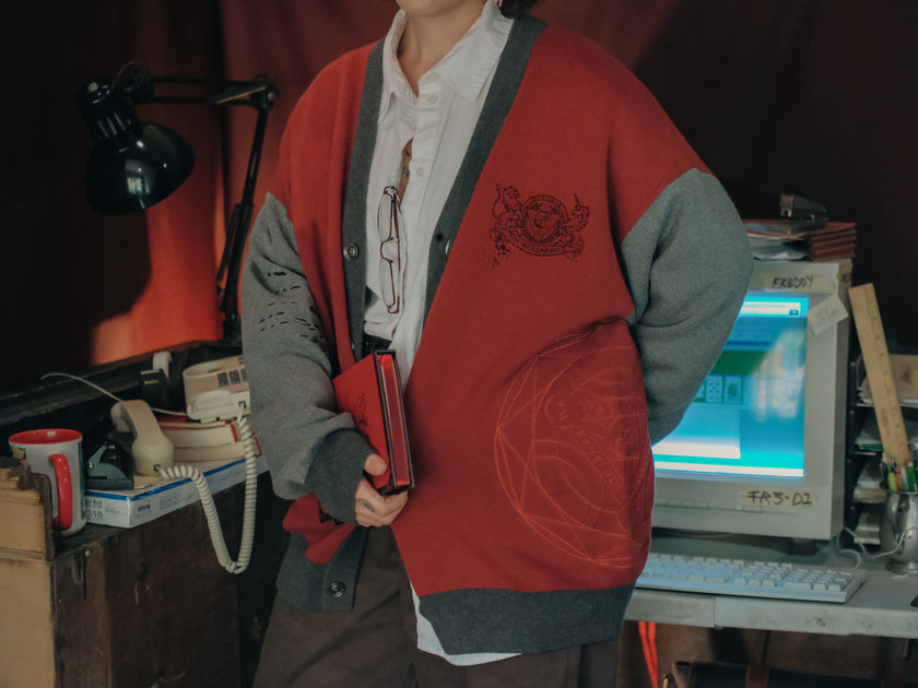 Magnus Protocol, oversized, knit-cardigan, in red, black, and shades of grey. The cardigan features embroidered details of reading glasses, fountain pen, OIAR crest, and reads â€œOffice of Incident Assessment & Responseâ€.