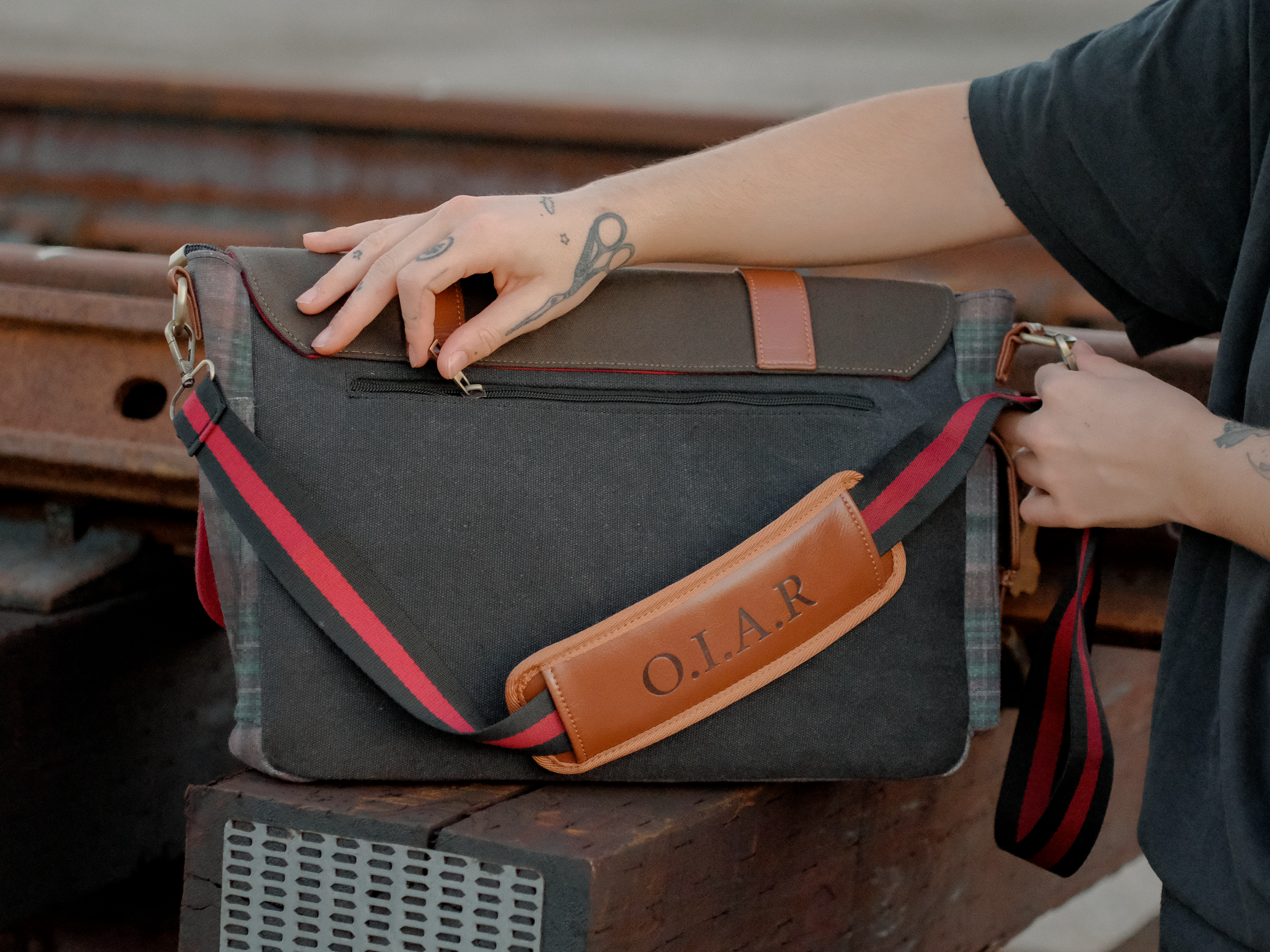 Knock Thrice x Rusty Quill canvas satchel with leather straps, plaid side panels, and an embroidered crest design. The bag is lined in plaid and includes a padded laptop pocket, adjustable cross-body strap, and magnetic closure.