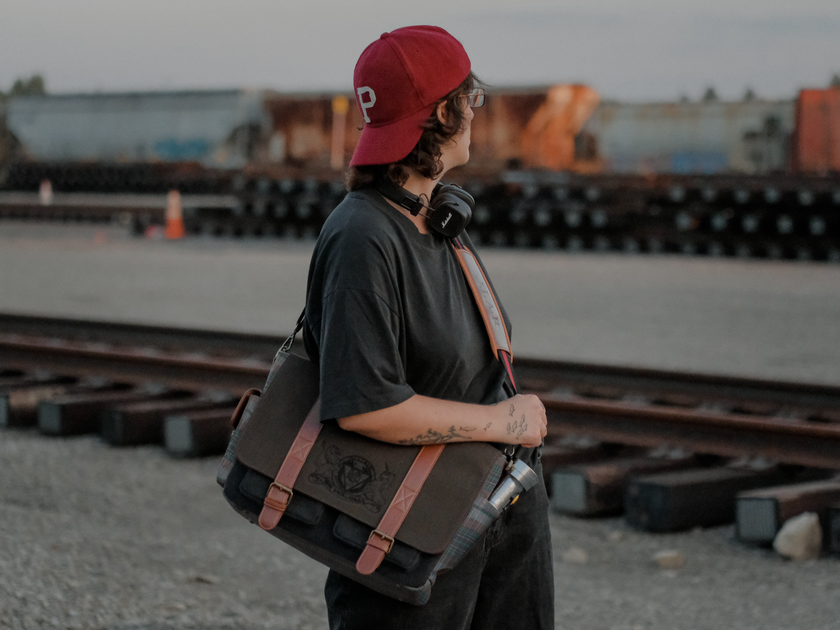 Knock Thrice x Rusty Quill canvas satchel with leather straps, plaid side panels, and an embroidered crest design. The bag is lined in plaid and includes a padded laptop pocket, adjustable cross-body strap, and magnetic closure.
