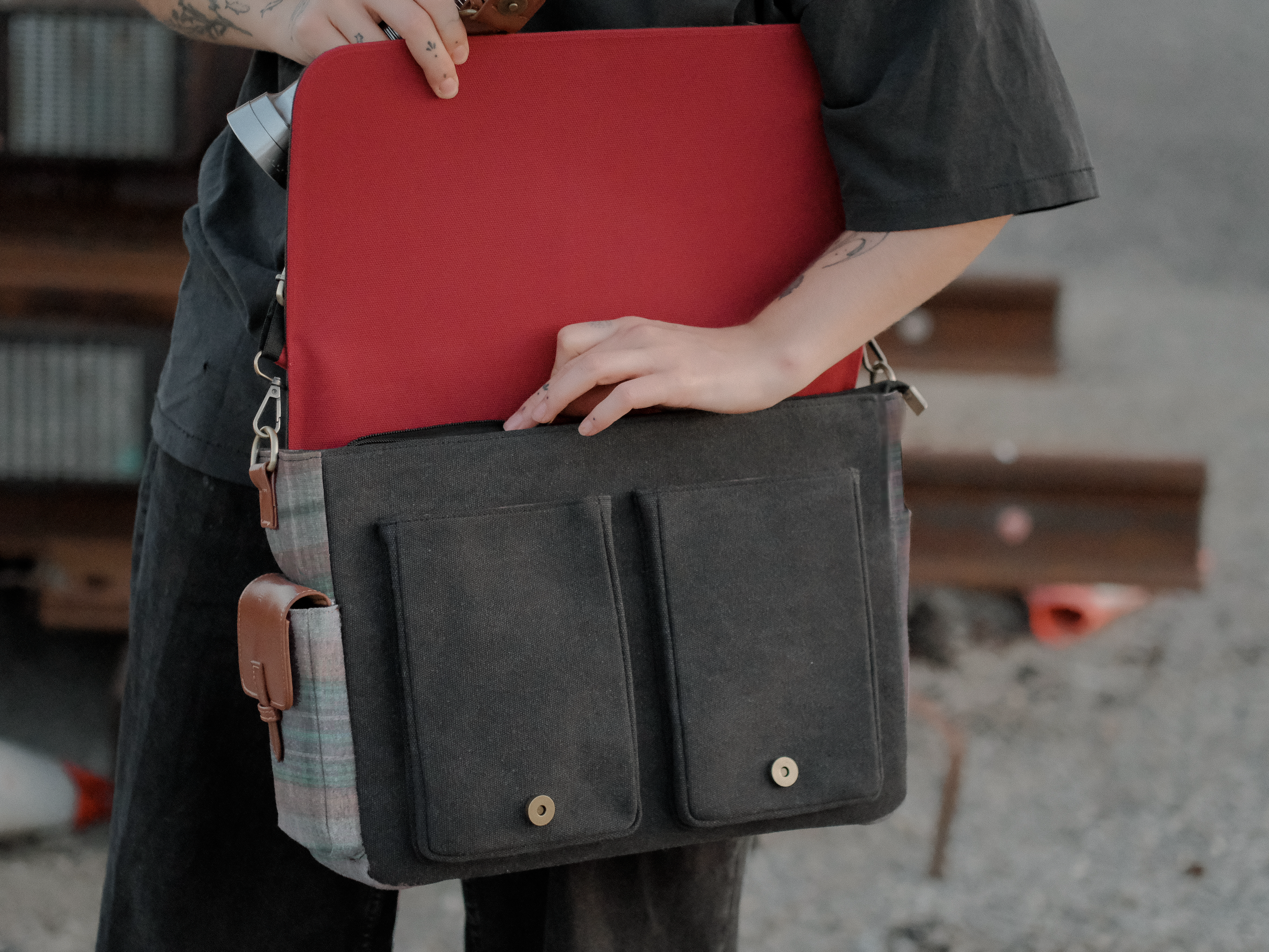 Knock Thrice x Rusty Quill canvas satchel with leather straps, plaid side panels, and an embroidered crest design. The bag is lined in plaid and includes a padded laptop pocket, adjustable cross-body strap, and magnetic closure.