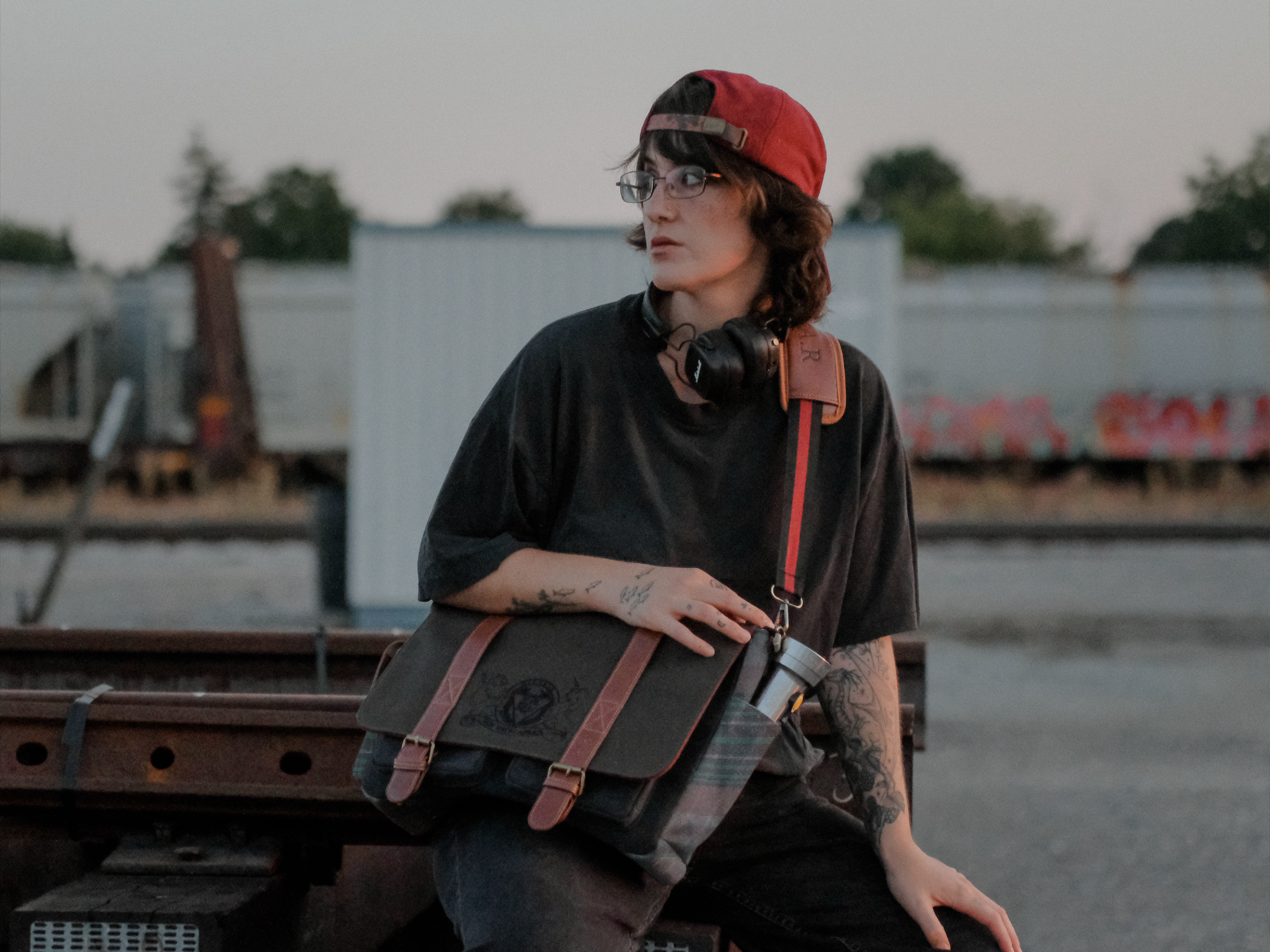 Knock Thrice x Rusty Quill canvas satchel with leather straps, plaid side panels, and an embroidered crest design. The bag is lined in plaid and includes a padded laptop pocket, adjustable cross-body strap, and magnetic closure.