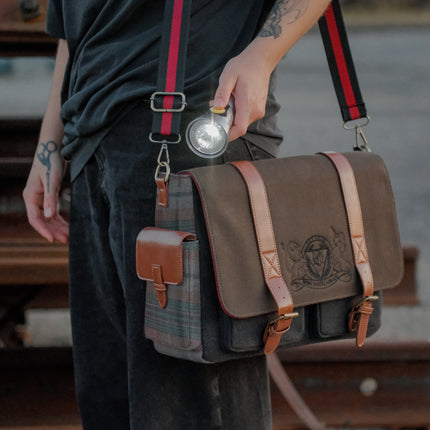 Knock Thrice x Rusty Quill canvas satchel with leather straps, plaid side panels, and an embroidered crest design. The bag is lined in plaid and includes a padded laptop pocket, adjustable cross-body strap, and magnetic closure.