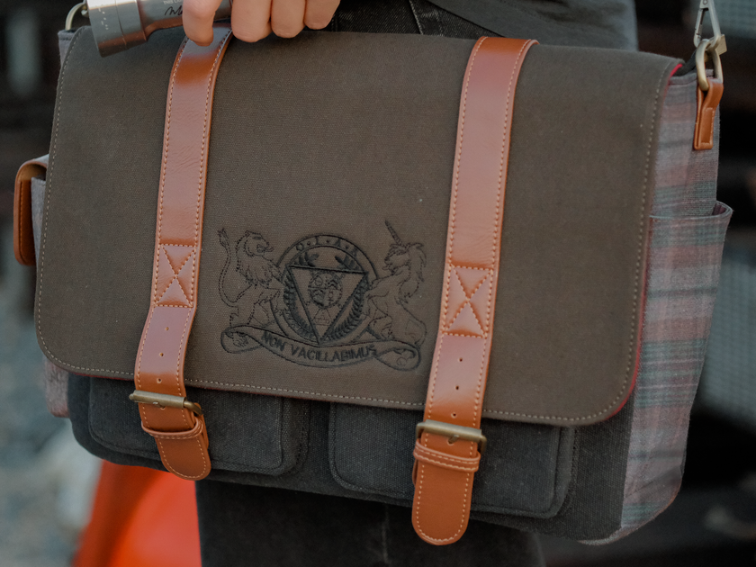 Knock Thrice x Rusty Quill canvas satchel with leather straps, plaid side panels, and an embroidered crest design. The bag is lined in plaid and includes a padded laptop pocket, adjustable cross-body strap, and magnetic closure.