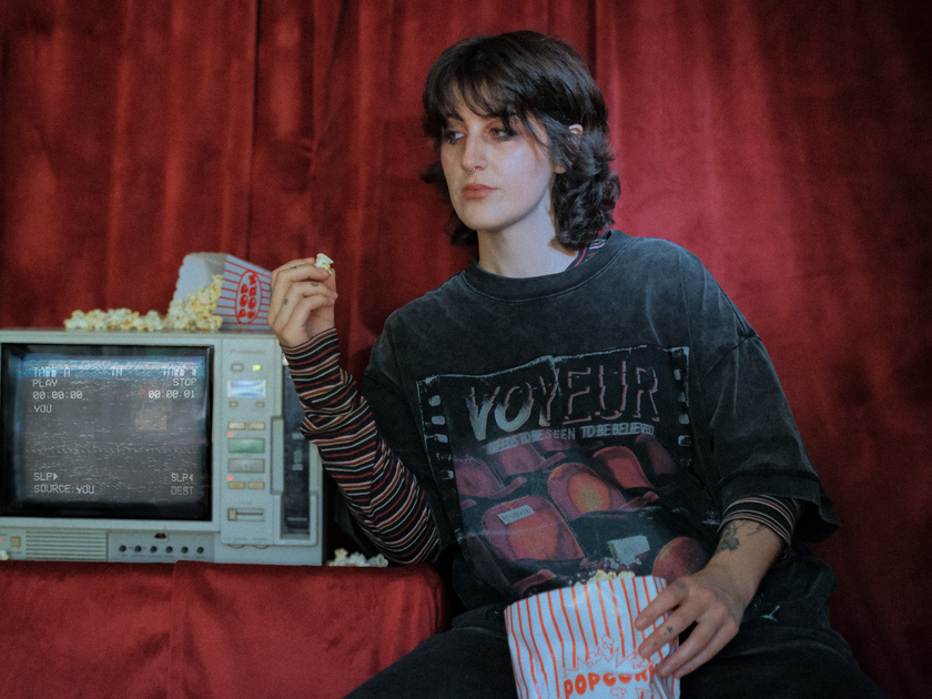 Knock Thrice x Rusty Quill, oversized, stone-washed cotton tee, with a graphic design titled 'Voyeur,' showcasing an eerie movie theatre with red seats, bold text reading 'Needs to be seen to be believed,' and film-inspired details.