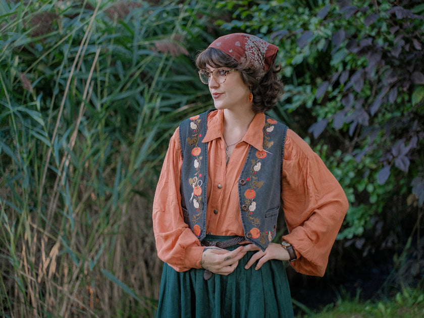 Cropped, vintage-style denim vest in a deep charcoal color, adorned with detailed embroidery of pumpkins, leaves, and vines in autumnal hues. The vest features a V-neckline, button closures, two front pockets, and intricate stitched details creating a rustic and whimsical aesthetic. 
