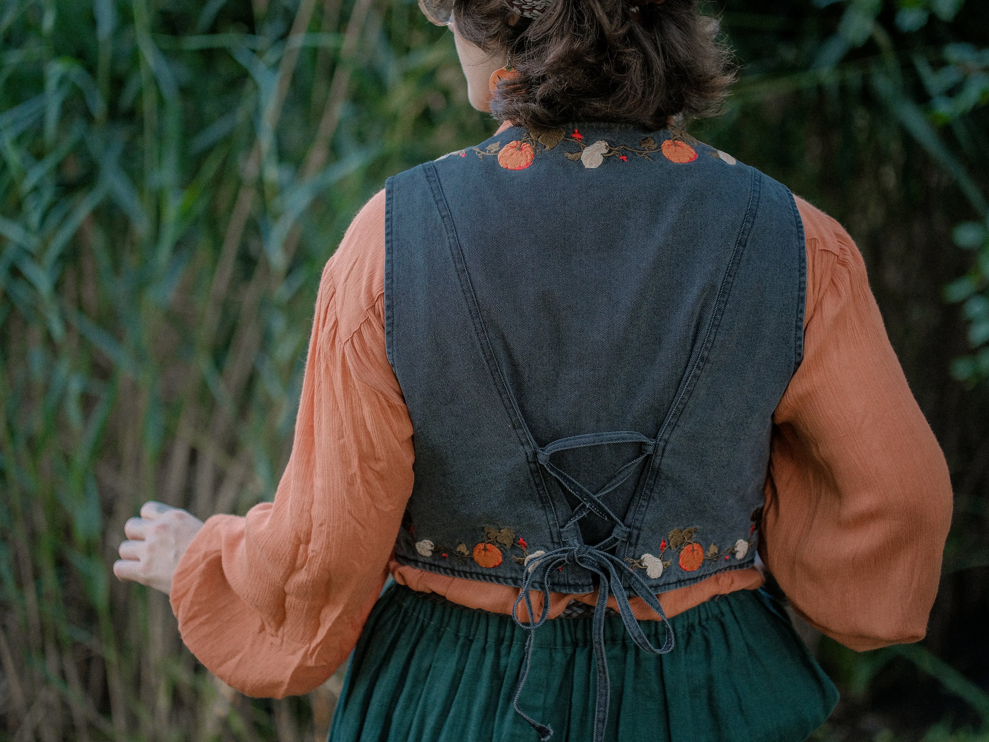 Cropped, vintage-style denim vest in a deep charcoal color, adorned with detailed embroidery of pumpkins, leaves, and vines in autumnal hues. The vest features a V-neckline, button closures, two front pockets, and intricate stitched details creating a rustic and whimsical aesthetic. 
