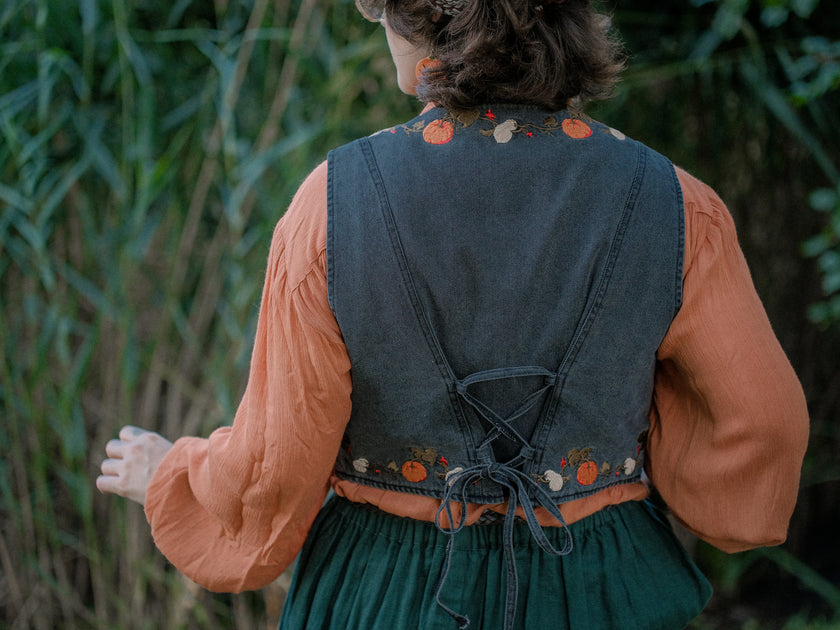 Cropped, vintage-style denim vest in a deep charcoal color, adorned with detailed embroidery of pumpkins, leaves, and vines in autumnal hues. The vest features a V-neckline, button closures, two front pockets, and intricate stitched details creating a rustic and whimsical aesthetic. 