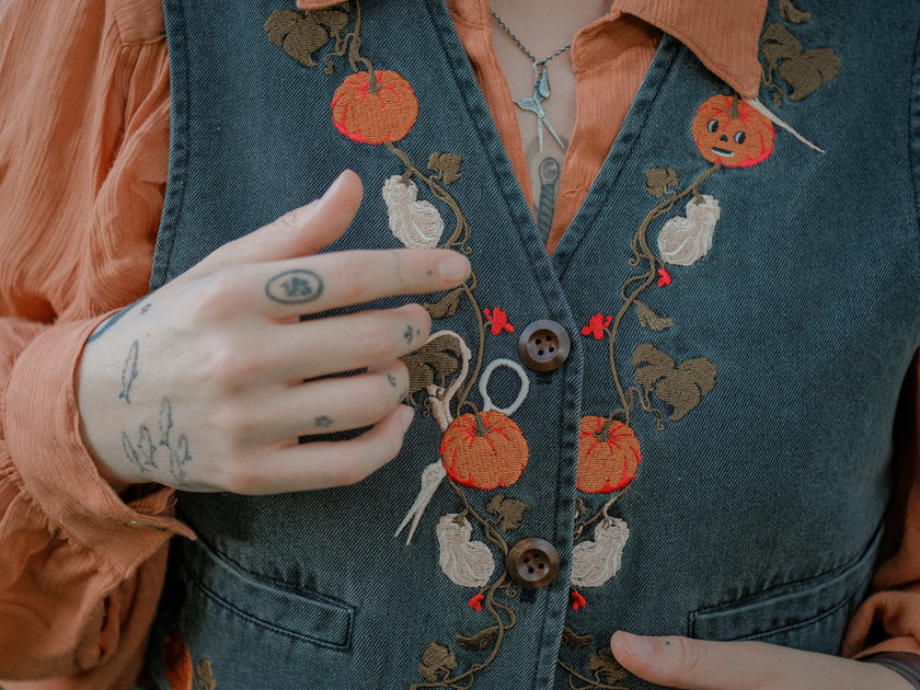 Cropped, vintage-style denim vest in a deep charcoal color, adorned with detailed embroidery of pumpkins, leaves, and vines in autumnal hues. The vest features a V-neckline, button closures, two front pockets, and intricate stitched details creating a rustic and whimsical aesthetic. 