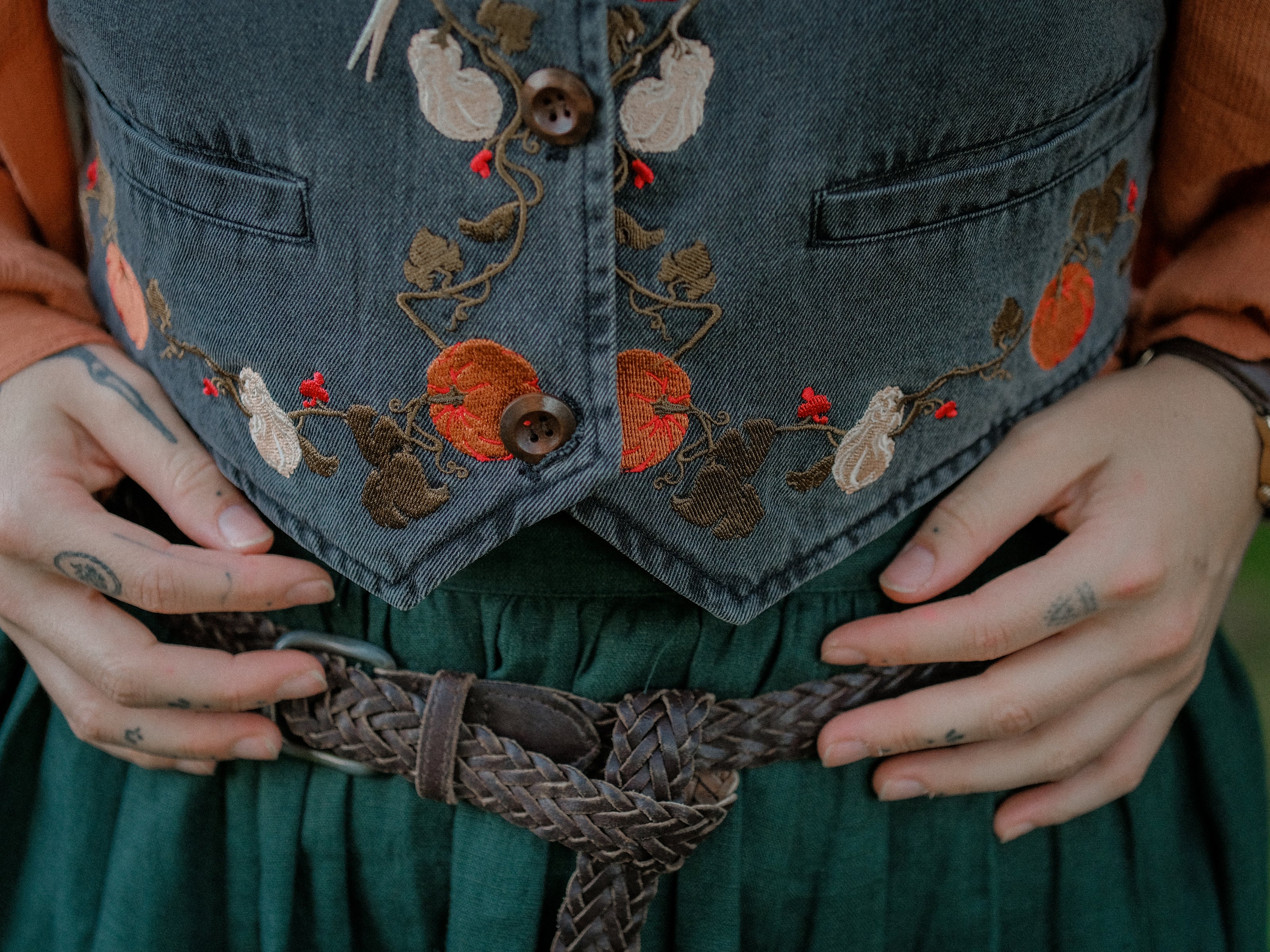 Cropped, vintage-style denim vest in a deep charcoal color, adorned with detailed embroidery of pumpkins, leaves, and vines in autumnal hues. The vest features a V-neckline, button closures, two front pockets, and intricate stitched details creating a rustic and whimsical aesthetic. 