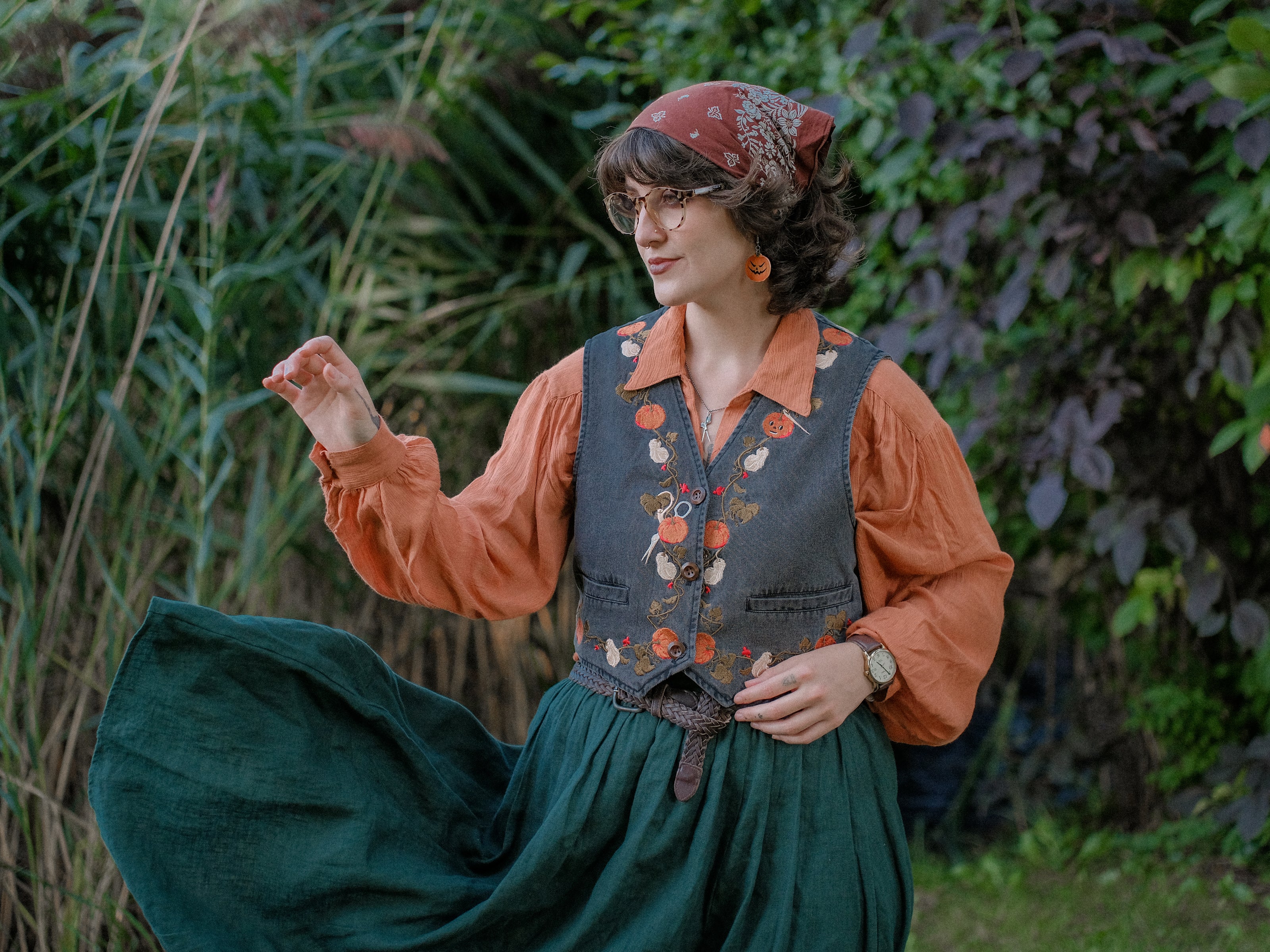 Cropped, vintage-style denim vest in a deep charcoal color, adorned with detailed embroidery of pumpkins, leaves, and vines in autumnal hues. The vest features a V-neckline, button closures, two front pockets, and intricate stitched details creating a rustic and whimsical aesthetic. 