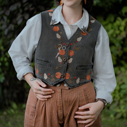 Cropped, vintage-style denim vest in a deep charcoal color, adorned with detailed embroidery of pumpkins, leaves, and vines in autumnal hues. The vest features a V-neckline, button closures, two front pockets, and intricate stitched details creating a rustic and whimsical aesthetic. 