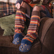 A person sitting on a brown couch wearing long striped over-the-knee socks in earthy tones, sheer black tights, and blue socks with gold stars at the foot.