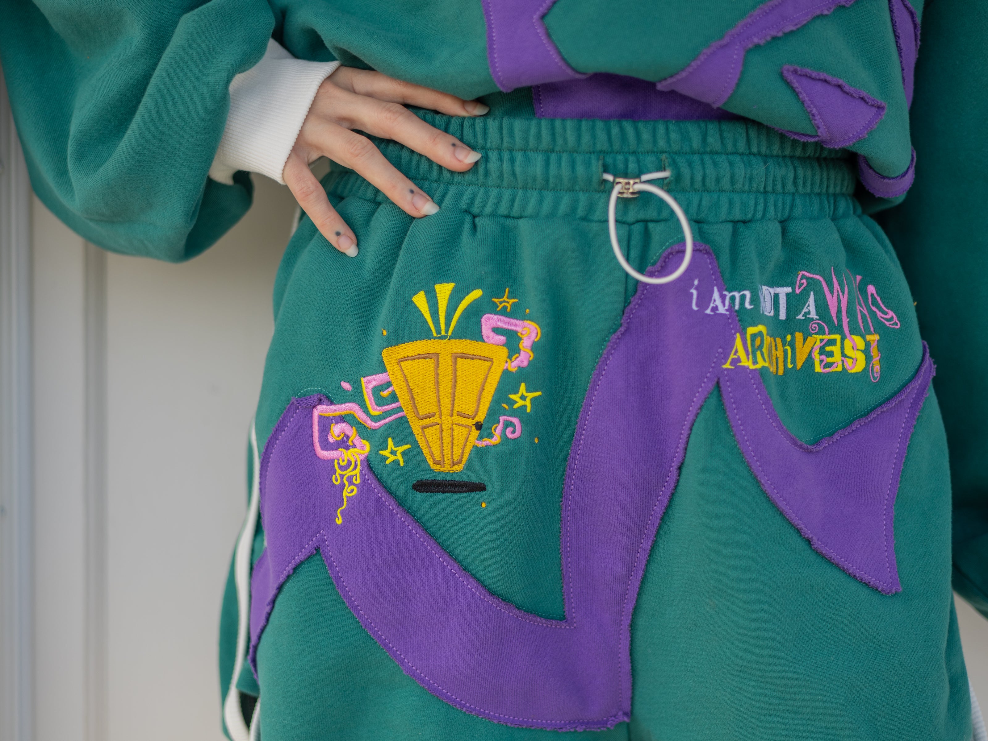 Magnus Archives x Knock Thrice “I’m a What” long shorts in green and purple, 100% cotton graphic shorts with embroidered details