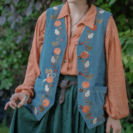 Vintage-style, blue light-wash denim vest, adorned with detailed embroidery of pumpkins, gourds, leaves, and vines in autumnal hues. The vest features a V-neckline, button closures, two front pockets, and intricate stitched details creating a rustic and whimsical aesthetic. 