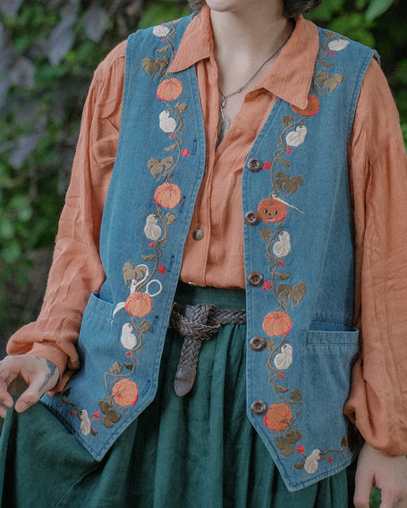 Vintage-style, blue light-wash denim vest, adorned with detailed embroidery of pumpkins, gourds, leaves, and vines in autumnal hues. The vest features a V-neckline, button closures, two front pockets, and intricate stitched details creating a rustic and whimsical aesthetic. 