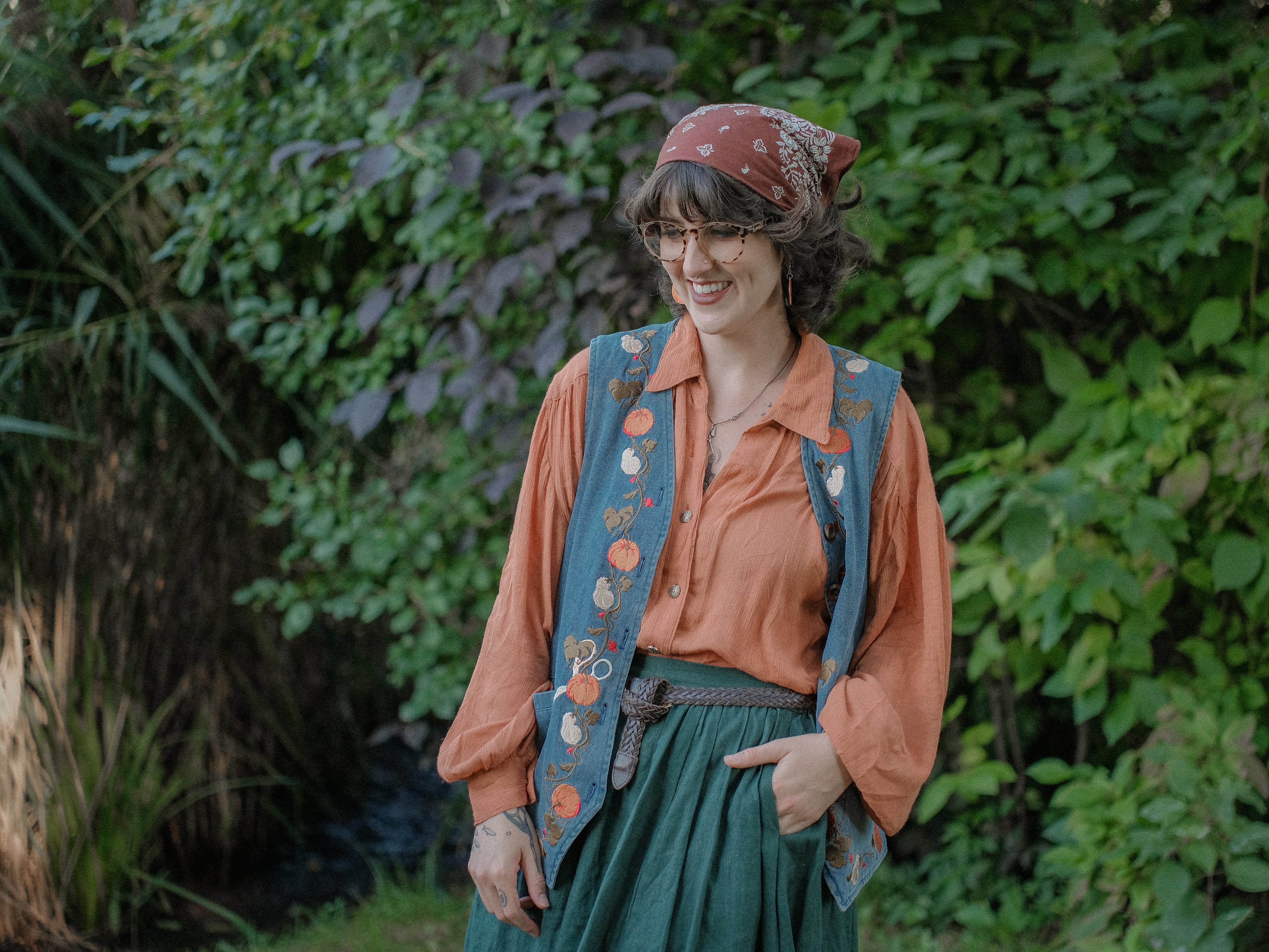 Vintage-style, blue light-wash denim vest, adorned with detailed embroidery of pumpkins, gourds, leaves, and vines in autumnal hues. The vest features a V-neckline, button closures, two front pockets, and intricate stitched details creating a rustic and whimsical aesthetic. 